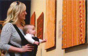 A blond woman holds a baby in a carrier looking at a colorful hung on the wall. 
