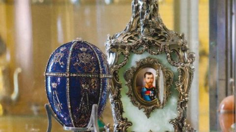 Ornate decorations, such as a bejeweled blue egg and a depiction of a Russian monarch, sit on a glass table