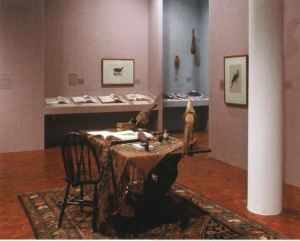 A table with one chair sit on an oriental rug in a gallery with fabric and two stuffed pheasants. 