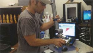 A man works with a small camera filming an artifact on a table with a computer behind him. 