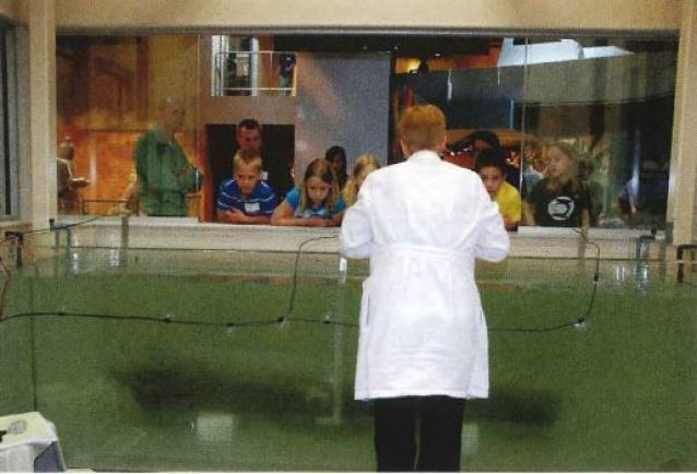 View of a woman in a white lab coat standing with her back to the camera facing a tank filled with water and something mirky inside while a group of children lean on the windowsill of an open window looking down into the tank. 