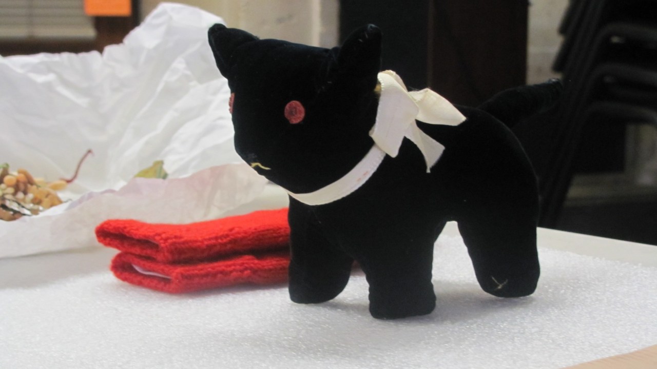 A small black stuffed animal cat sits on a table with various other collections items behind it.