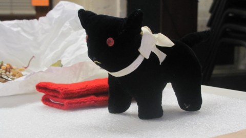 A small black stuffed animal cat sits on a table with various other collections items behind it.