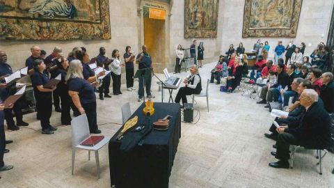 Multiple people in hospital scrubs sing while one woman sit in a chair facing them playing guitar. many people sit behind the woman watching the performance.
