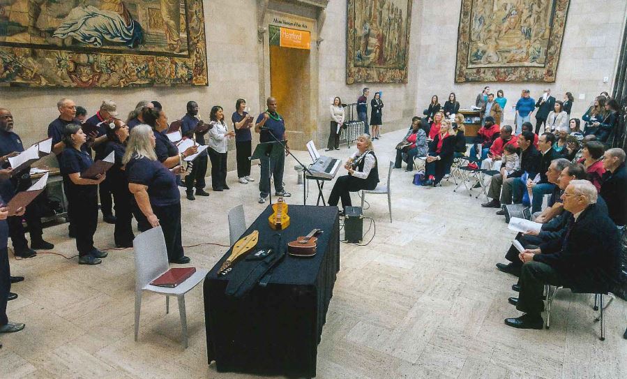 Multiple people in hospital scrubs sing while one woman sit in a chair facing them playing guitar. many people sit behind the woman watching the performance.