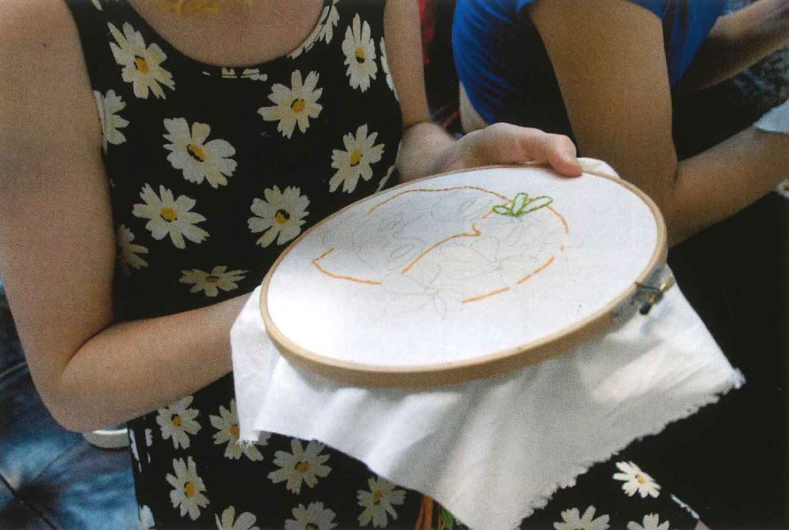 A woman in a flower dress embroiders the outline of a woman onto a white cloth.