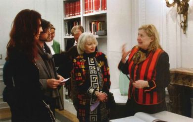 Several people listen to a docent while standing in a room with bookshelves. 