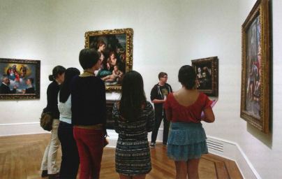 Several people listen to a tour guide explain a painting on a wall with several other paintings in an octagonal shaped room. 