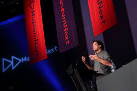 A male speaker stands behind a podium on stage with several red and black banners that read Museum Next hanging above his head.