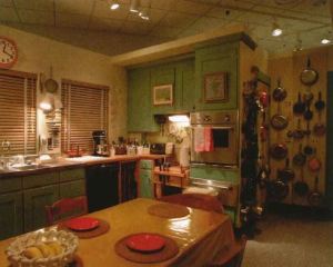 View of the interior of Julia Child's kitchen with green painted cabinets and a couple of paintings hung on the cabinet doors, two windows with wooden blinds pulled shut, a long rectangular table in the center and a lot of pots and pans hung on a wall behind the cabinet wall. 