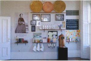 View looking directly at an exhibition wall with various shelves arranged with items above which are hung images, copper trays, and a text panel. 