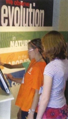Image showing two girls looking at a computer interactive with the wall panel behind them "We observe evolution"