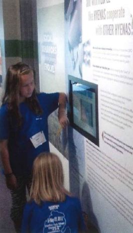 Two girls looking at a wall text with a computer screen inserted into it.