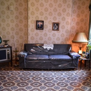 View of the interior of one of the Tenement Museums exhibits with a couch covered in clear plastic sitting on a patterned rug with a side table and lamp to the right and a 1960s television to the left. The walls have a patterned wallpaper. 