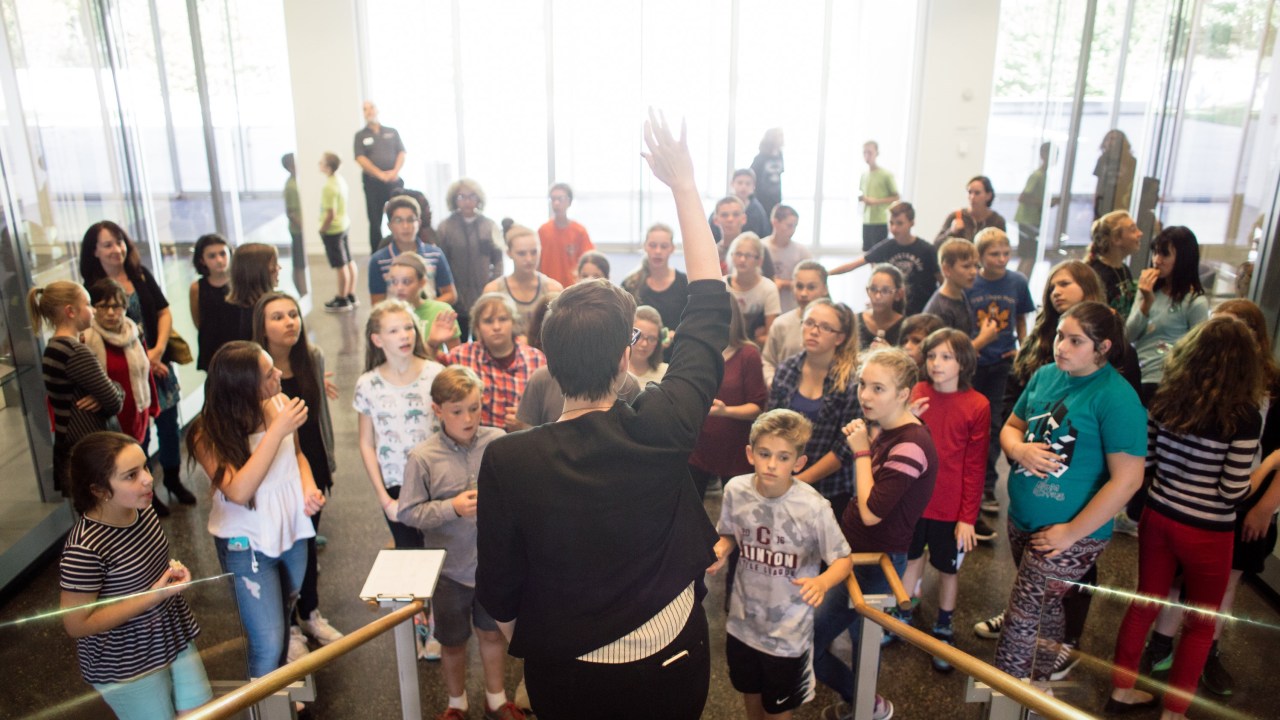 A large group of children stand in front of a woman who has her back to the camera.
