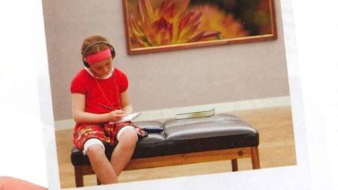 Image of a person's hand holding a Poloroid image of a girl sitting on a bench in a gallery.
