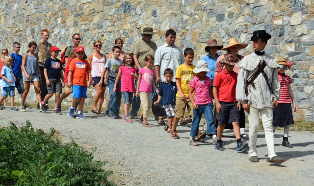 A double line of children and adults walking next to a stone wall.