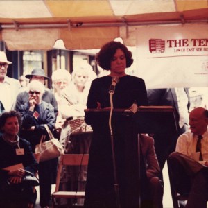 View of a woman standing at a podium with various people in the background.