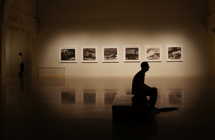 A sepia-toned image of a man sitting in a gallery contemplating a work of art. The wall behind him is backlit with a series of photographs.
