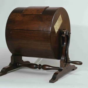 A wooden structure with a round barrel situated on top of a trestle with a wooden crank and an opening in the top of the barrel