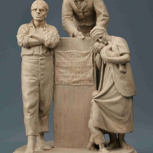 White marble sculpture with two men standing in front of and behind an auction block with a woman standing to the right hand side carrying a baby that she is kissing in a longing way.