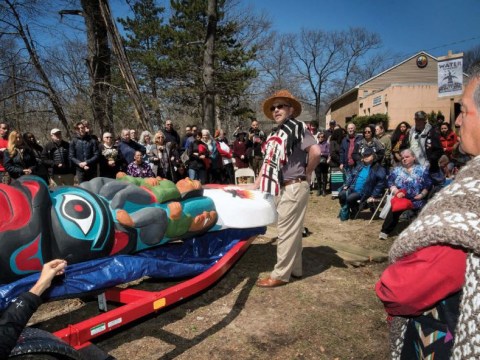 A group of people stand watching a two native men start to carry a colorful totum pole laying down off a trailer is wheeled past.