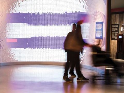 A couple of blurred people interact with a museum interactive with a shimmery glass wall behind them.