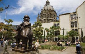 Exterior shot of the plaza with a sculpture to the left, a man emerging from behind the sculpture and an ornately designed domed structure looms in the background.