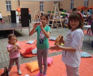 Three young girls play instruments standing on a mat with various multi-colored pillows lying around. 