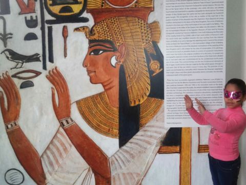 A young girl mimicks the Egyptian hygroglyph depicted in a large wall panel in a museum exhibition.