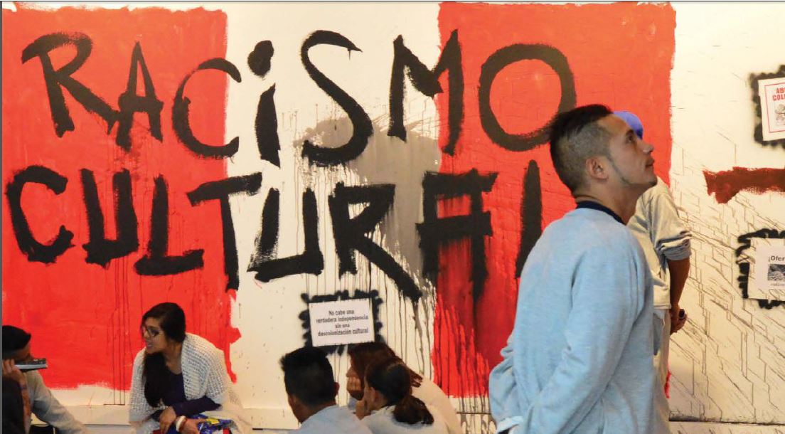 A group of people sit on the floor an done young hispanic man looks at a wall panel inside the museum's exhibition.
