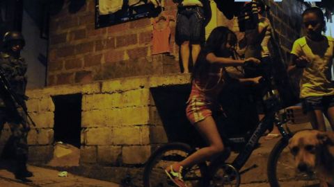 Two young girls ride their bicycles on a sidewalk next to a cinderblock house with a uniformed man with a large rifle behind them.