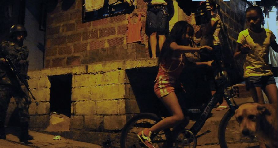 Two young girls ride their bicycles on a sidewalk next to a cinderblock house with a uniformed man with a large rifle behind them.