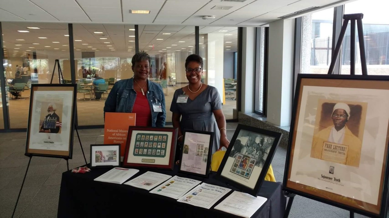 Two black women stand behind a table with various framed stamp collections.