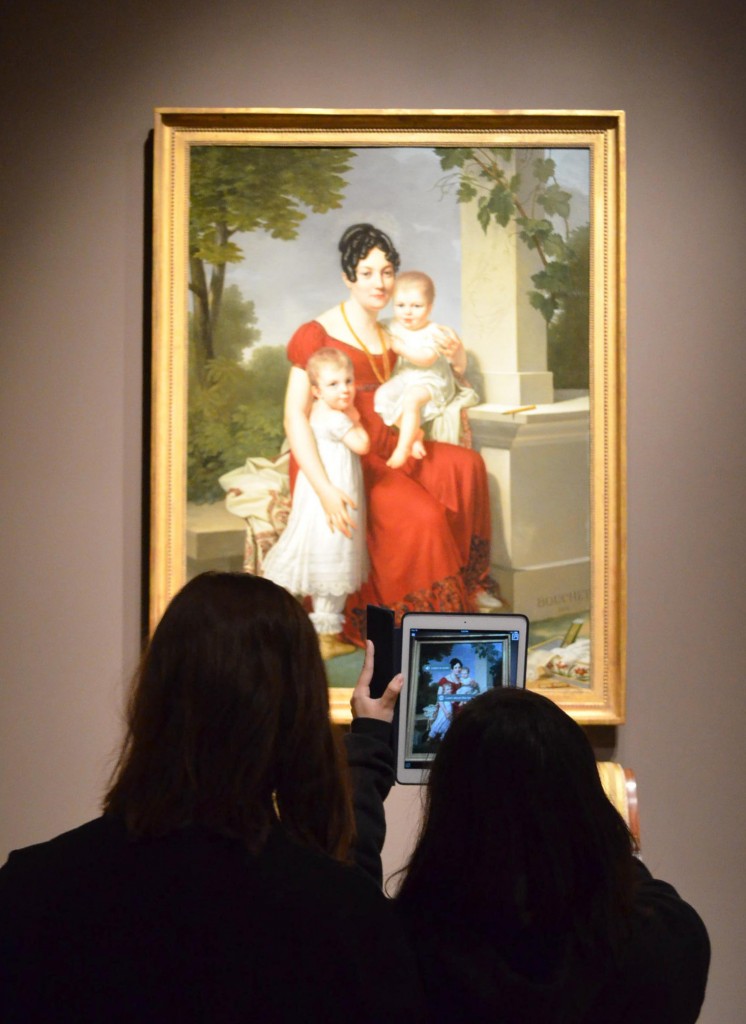 Two teens face a framed painting of a mother with two children on her lap. The two teens hold up a cell phone that is displaying the painting with additional text.