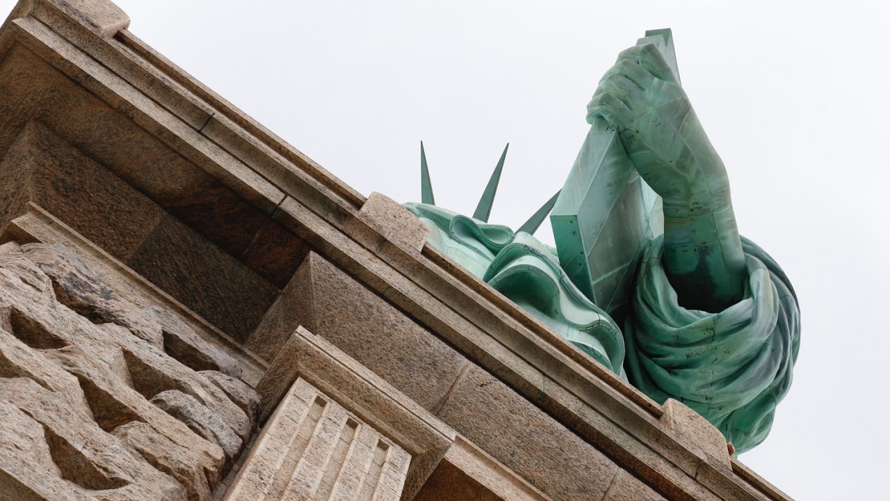 View of the Statue of Liberty taken from below at an angle so that all that is visible is her hand with the book in it an da few spikes from her crown an dthe edge of the plinth on which she stands.