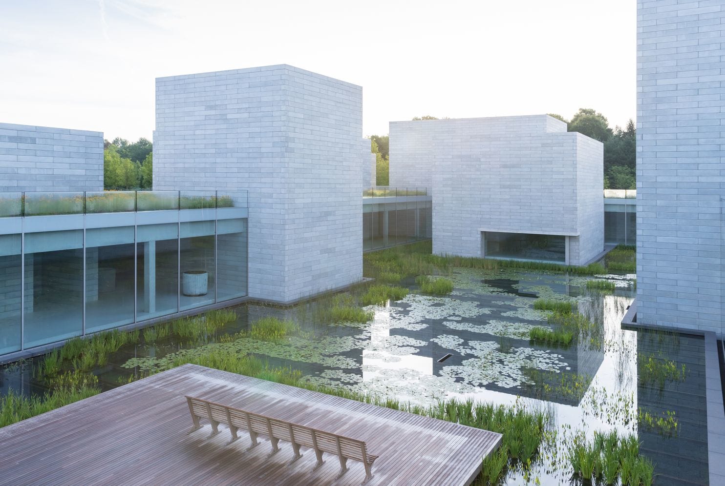 A pond surrounded by modern, grey stone buildings. A bench is facing the pond.