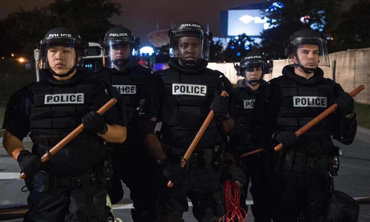 Five police officers in full riot gear lined up across a city street at night.