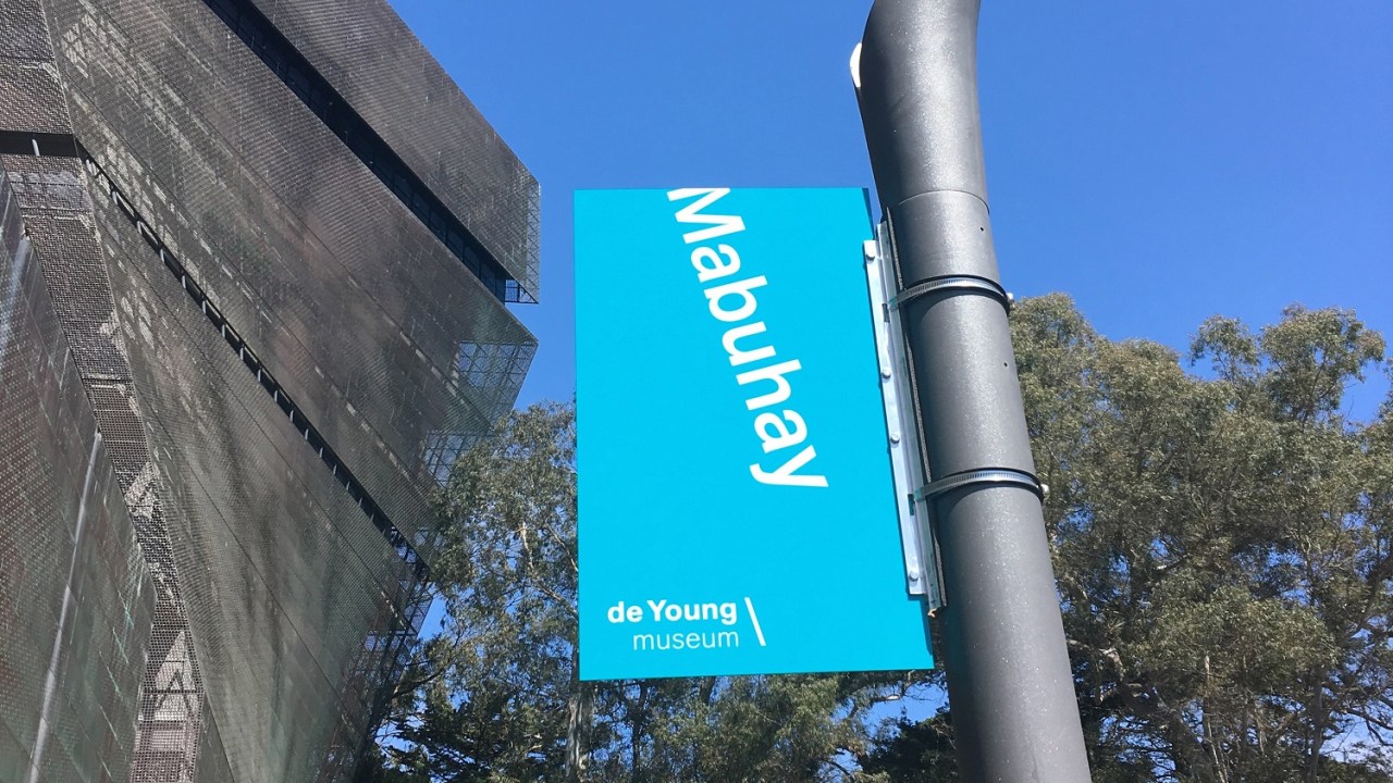 Exterior image of the de Young museum with a Mabuhay/Welcome banner in teal with white letters hanging from a lamp post.