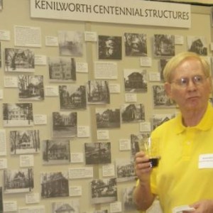 A white man in a yellow collared short sleeve shirt wearing a nametag stands in front and to the right of a wall with the words Kenilworth Centennial Structures and various black and white pictures mounted on it. 