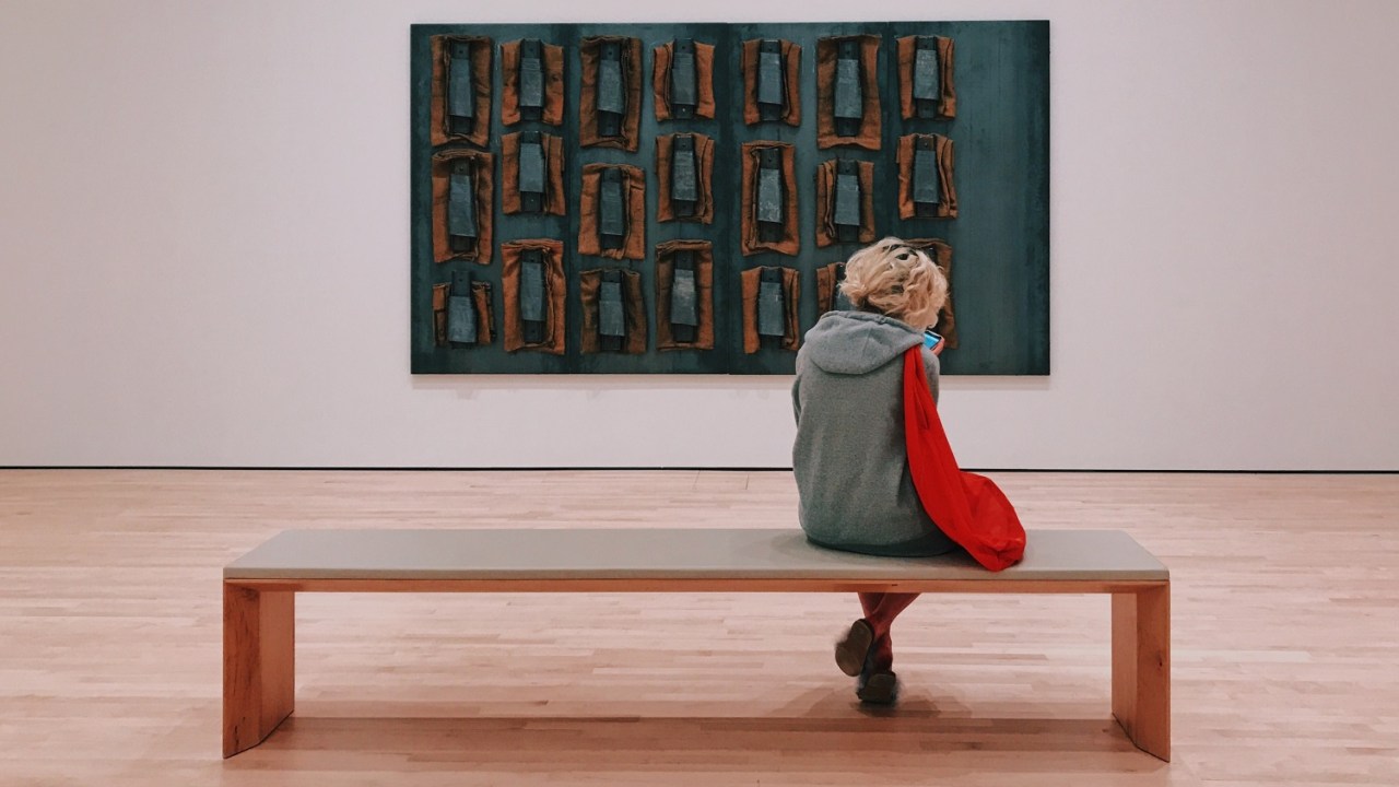 A woman sits on a long backless bench in front of a contemporary art piece.