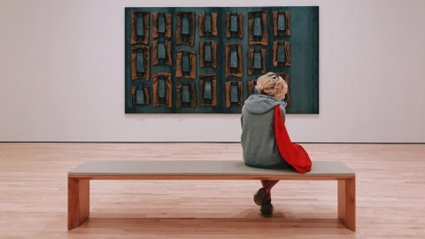 A woman sits on a long backless bench in front of a contemporary art piece.