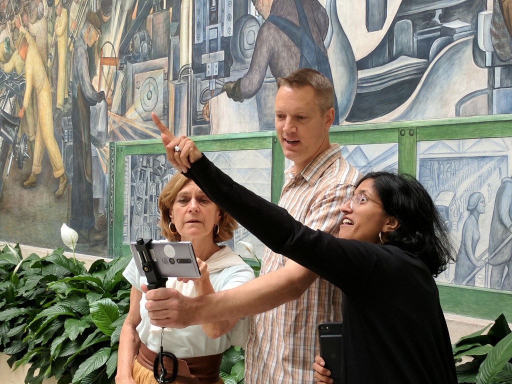 A white man stands in between one white woman and one brown woman holding a cell phone on a holster out in front of him, one of the women points to an object out of view. 