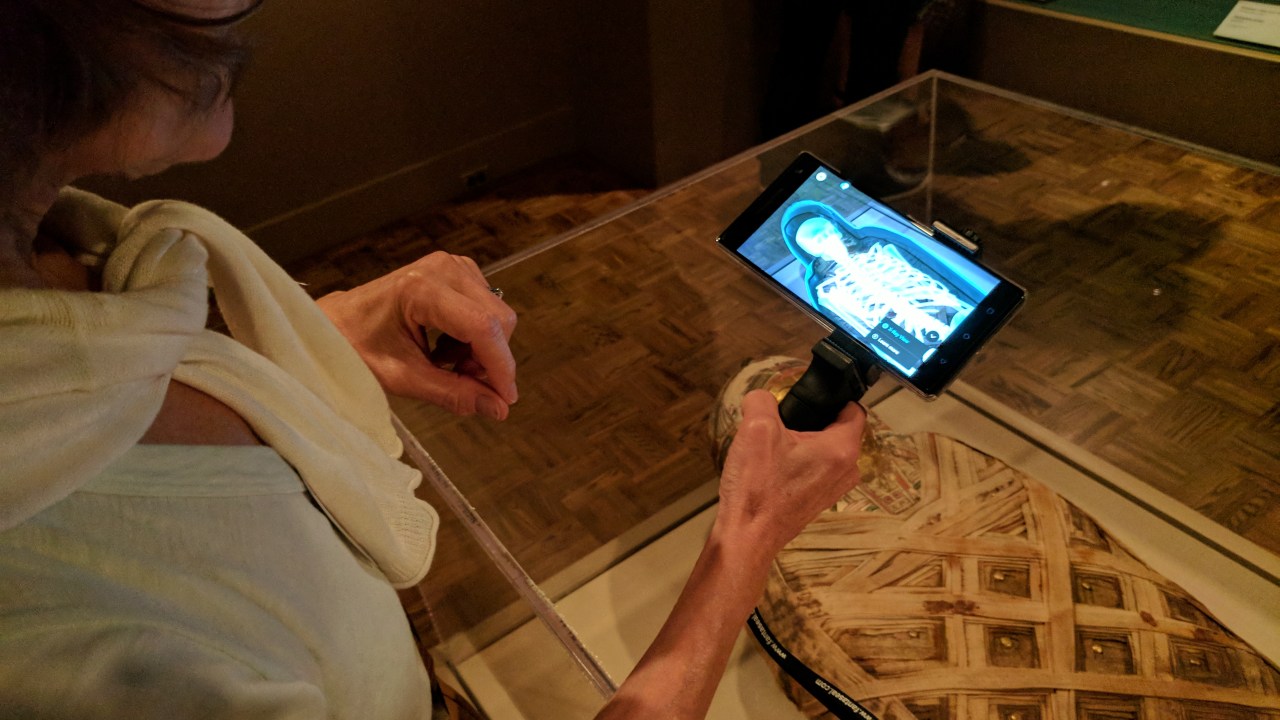 A white woman holds a phone holster in her right hand showing an x-ray of a mummy as she stands at a vitrine with a mummy displayed inside.