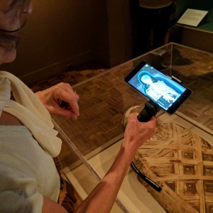 A white woman holds a phone holster in her right hand showing an x-ray of a mummy as she stands at a vitrine with a mummy displayed inside. 