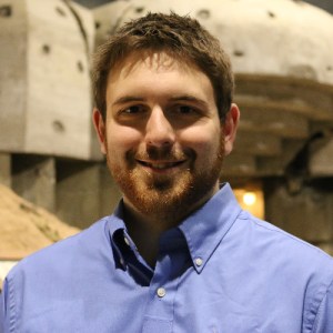 A young white man smiles at the camera with a scruffy beard and wearing a blue button down shirt.