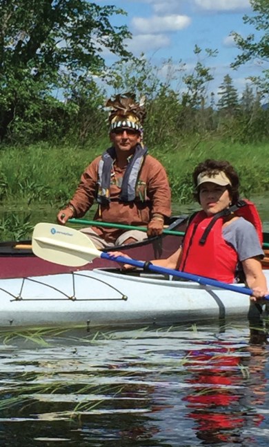 A man sits in a canoe with a feathered headress on and a woman sits in a separate canoe wearing a tan visor and life vest.