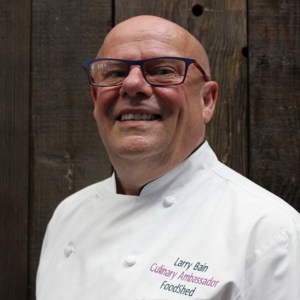 A bald white man stands in a chef 's white jacket in front of a wooden wall smiling broadly at the camera wearing glasses.