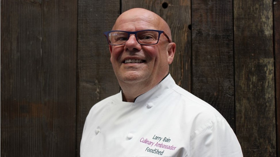 A bald white man stands in a chef 's white jacket in front of a wooden wall smiling broadly at the camera wearing glasses.