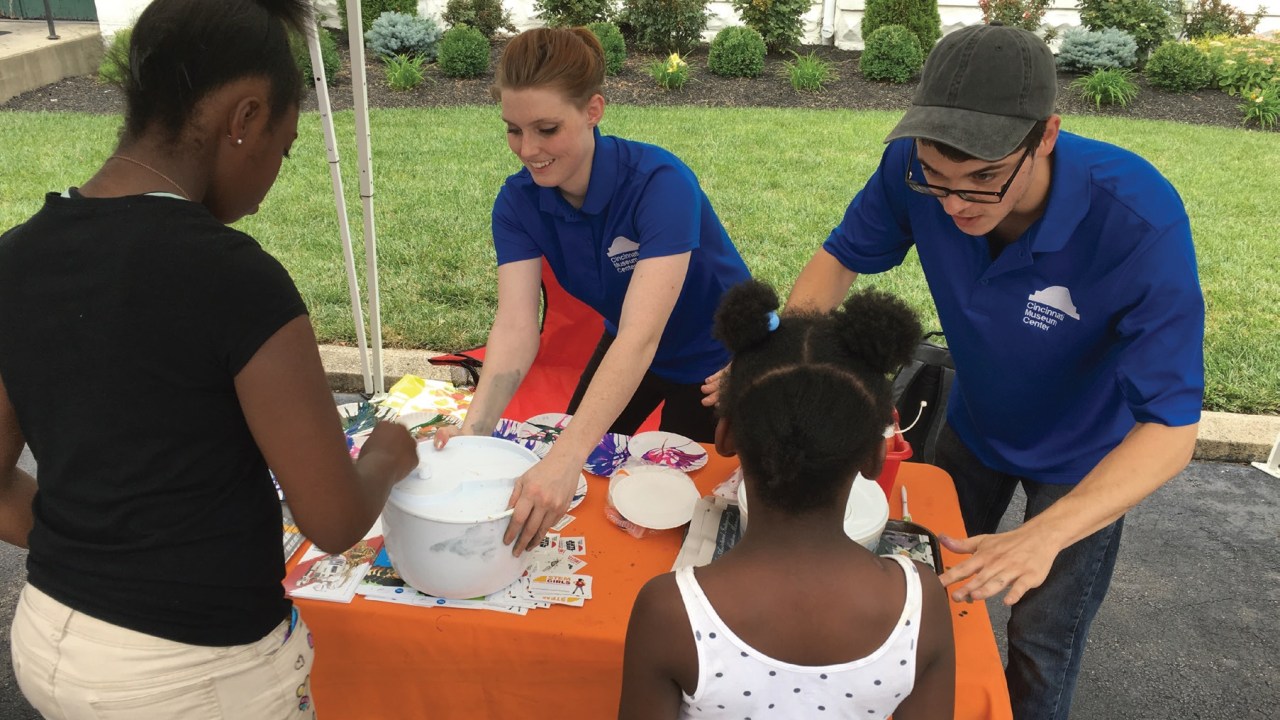 Two (male and female) white people in blue shirts help two (both female, one adult, one child) black people with a spin art presentation.